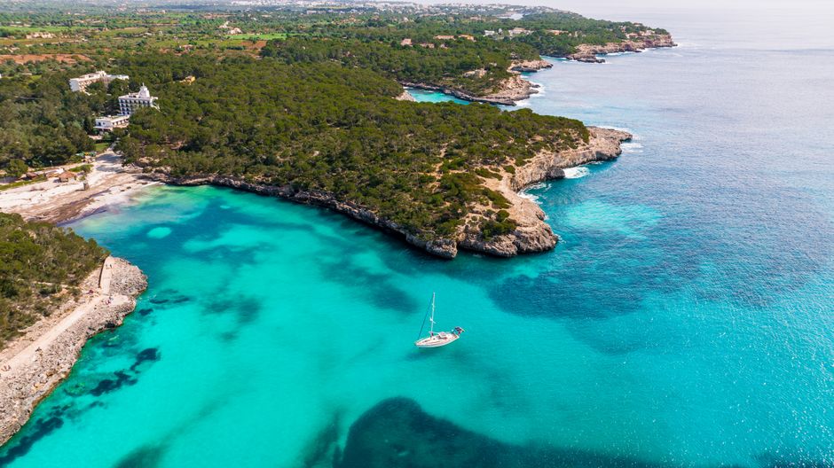 Breathtaking aerial view of a turquoise cove in Mallorca, Spain, featuring a lone sailboat.